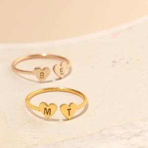 May include: Two gold rings with heart-shaped charms. The charms are engraved with the letters "B" and "E" on one ring and "M" and "T" on the other.