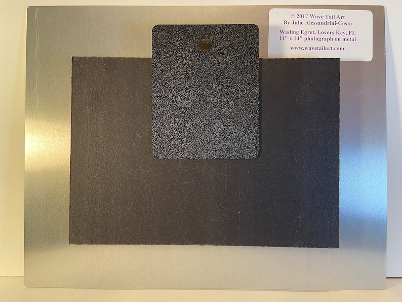 May include: A metal photograph with a silver border and a black backing. The photograph is labeled "Wading Egret, Lovers Key, FL" and measures 11 x 14 inches. The back has a black textured rectangle with a hole for hanging. The artist's name and website are also visible.
