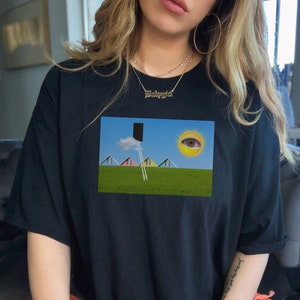 May include: Black t-shirt featuring a surreal landscape print. The graphic includes a ladder leading to a black rectangle, a sun with an eye, and colorful triangular shapes on a green field. The shirt is a casual, oversized style.
