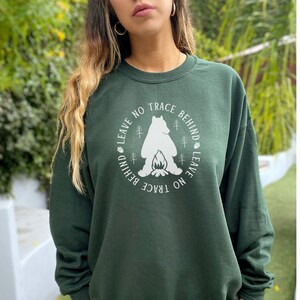 May include: A dark green sweatshirt with a white graphic of a bear sitting by a campfire. The graphic is surrounded by the text "Leave No Trace Behind Leave No Trace Behind".