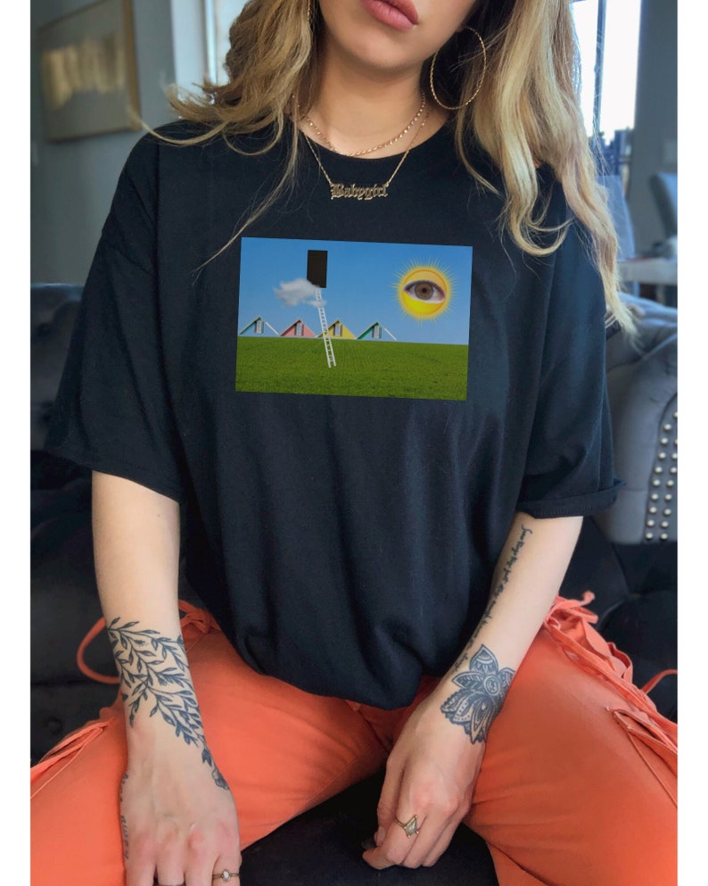 May include: Black t-shirt featuring a surreal landscape print. The graphic includes a ladder leading to a black rectangle, a sun with an eye, and colorful triangular shapes on a green field. The shirt is a casual, oversized style.