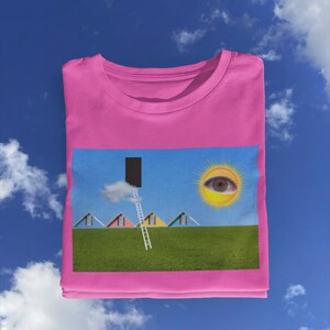 May include: A pink t-shirt featuring a surreal landscape design. The graphic includes colorful houses, a ladder leading to a black doorway, a sun with an eye, and a green field under a blue sky with clouds.