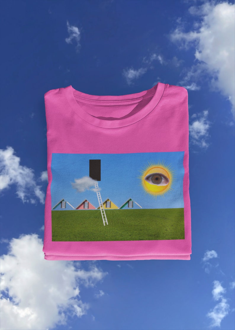 May include: A pink t-shirt featuring a surreal landscape design. The graphic includes colorful houses, a ladder leading to a black doorway, a sun with an eye, and a green field under a blue sky with clouds.