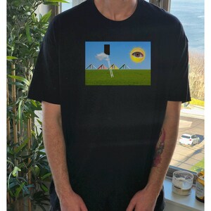 May include: Black t-shirt featuring a surreal landscape print. The print depicts a green field, a ladder leading to a black rectangle, a cloud, a sun with an eye, and colorful geometric shapes. The shirt has short sleeves and a crew neck.