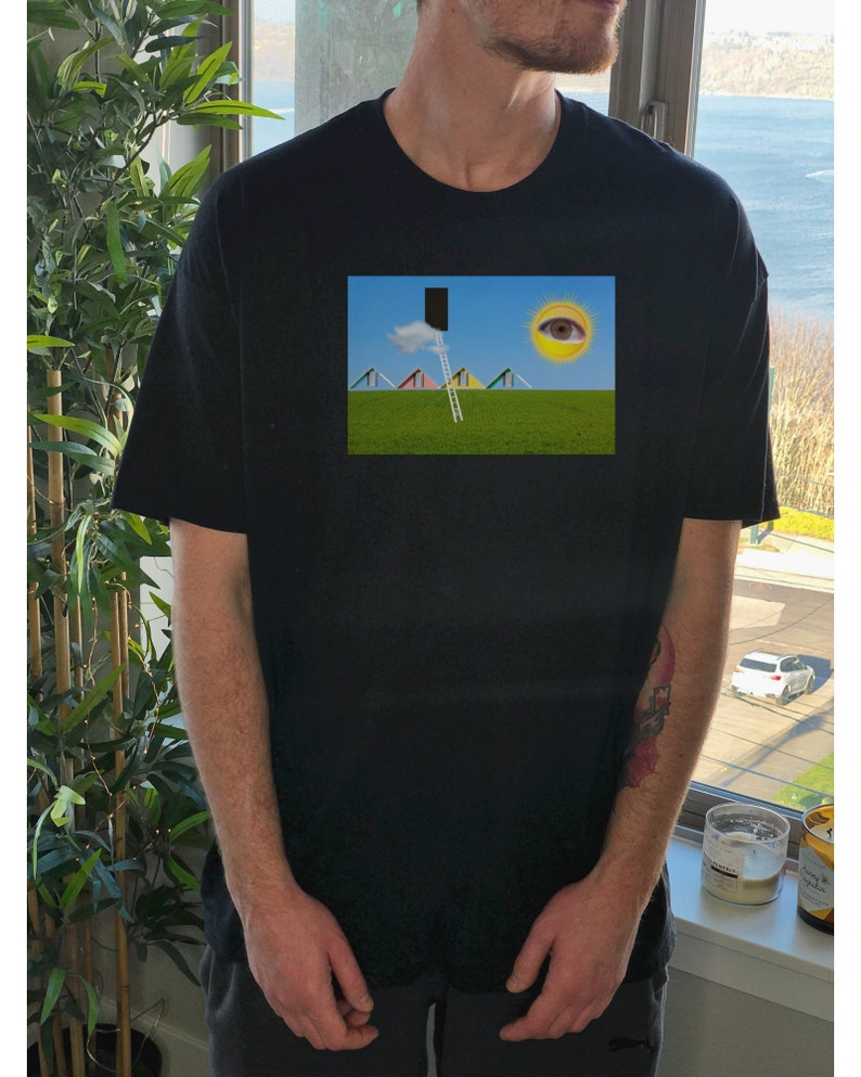 May include: Black t-shirt featuring a surreal landscape print. The print depicts a green field, a ladder leading to a black rectangle, a cloud, a sun with an eye, and colorful geometric shapes. The shirt has short sleeves and a crew neck.
