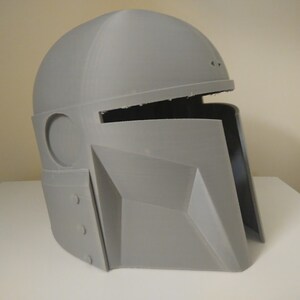 May include: A grey 3D printed helmet with a visor and a round opening on the side. The helmet is made of plastic and has a textured surface.