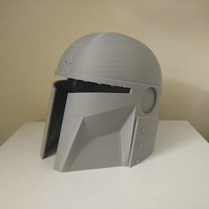 May include: A grey plastic helmet with a visor and a textured surface. The helmet has a round top and a pointed chin.