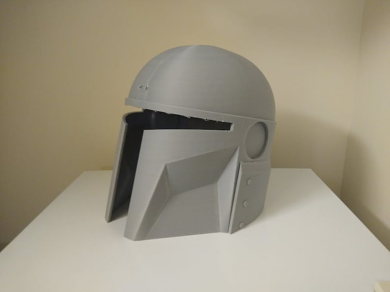 May include: A grey plastic helmet with a visor and a textured surface. The helmet has a round top and a pointed chin.