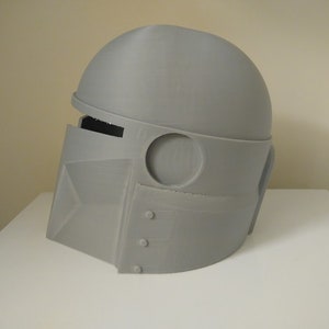 May include: A grey 3D printed helmet with a visor and a round top. The helmet has a circular opening on the side and a rectangular opening on the front.