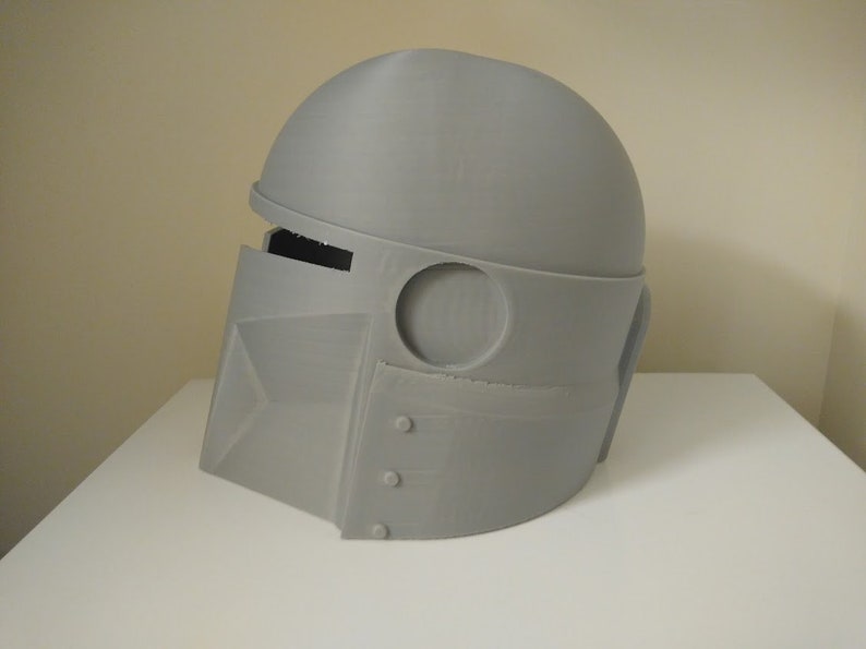 May include: A grey 3D printed helmet with a visor and a round top. The helmet has a circular opening on the side and a rectangular opening on the front.
