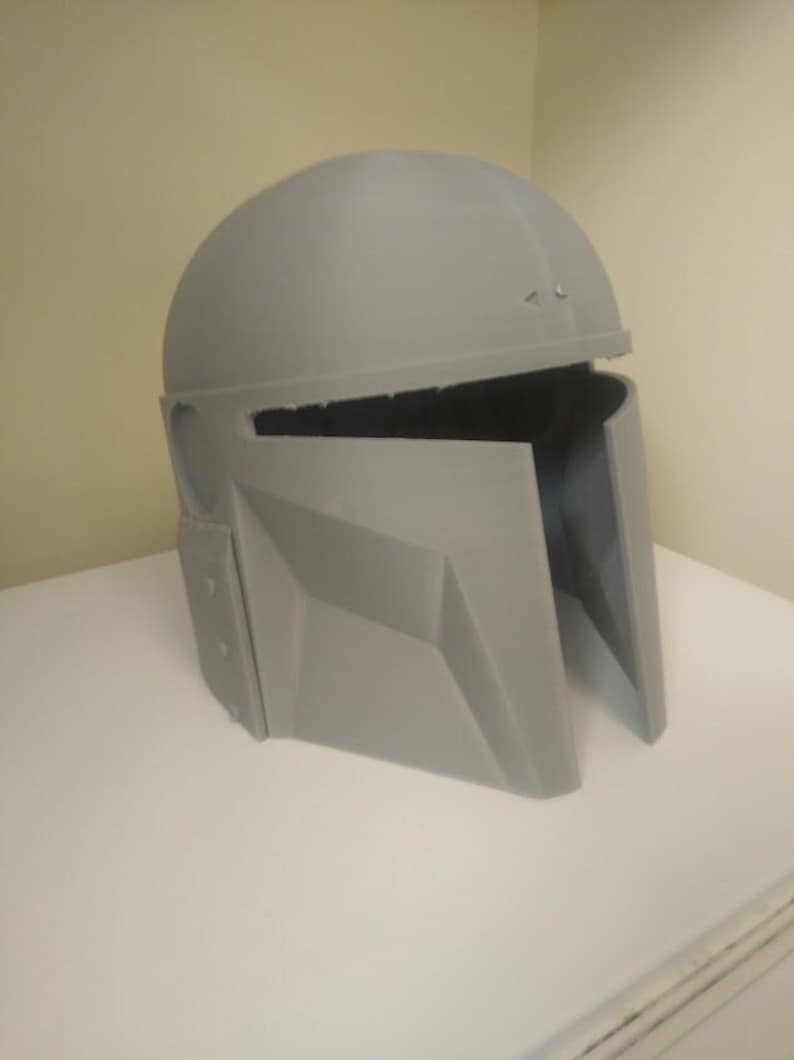 May include: A grey plastic helmet with a visor and a textured surface. The helmet is in two pieces, with the top piece removed.