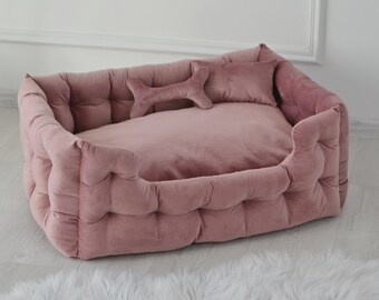 May include: A plush pink dog bed with a tufted design and a bone-shaped pillow. The bed is on a white fluffy rug.
