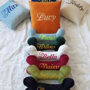 May include: A set of 9 personalized dog bone pillows. The pillows are in various colors and feature embroidered names, including Max, Lucy, Teddy, Luna, Willow, Bella, Daisy, Charlie, and Cooper.