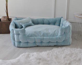 May include: A light blue dog bed with a tufted design and a matching pillow. The bed is filled with soft padding and has a raised edge for comfort.