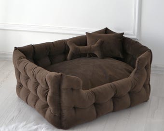 May include: A brown, tufted dog bed with a rectangular shape. The bed has a plush, cushioned design with a raised edge. Two small pillows, one bone-shaped and one square, are inside the bed. The bed is on a white, fluffy rug.