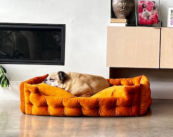 Op de afbeelding: Een oranje fluwelen hondenbed met een knoop-getuft ontwerp. Een slapende bulldog rust comfortabel in het bed. Het bed heeft een rechthoekige vorm en staat op een gepolijste betonnen vloer.
