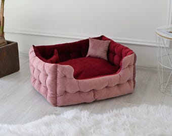May include: A plush dog bed with a burgundy and pink color scheme. The bed has a raised rim and a soft, quilted surface.