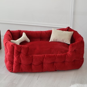 May include: A red dog bed with a tufted design and a white pillow. The bed has a raised edge and a bone-shaped pillow.