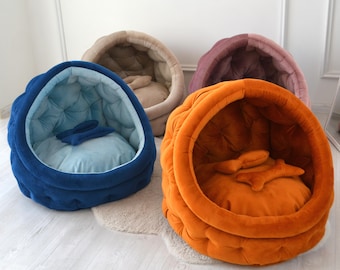 May include: Four pet beds in different colours, including blue, orange, beige, and purple. Each bed is shaped like a dome with a soft, plush interior.