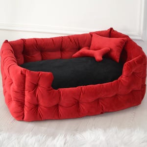 May include: A red, tufted dog bed with a black cushion. The bed has a rectangular shape with rounded edges and a low front. Two red pillows, one rectangular and one bone-shaped, are inside the bed.