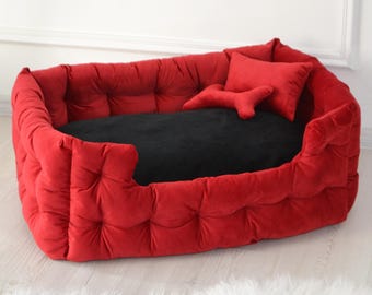 May include: A red, tufted dog bed with a black cushion. The bed has a rectangular shape with rounded edges and a low front. Two red pillows, one rectangular and one bone-shaped, are inside the bed.