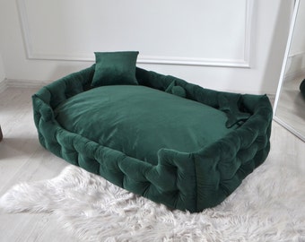May include: A deep green velvet dog bed with a tufted border and a matching pillow. The bed is on a white fluffy rug.