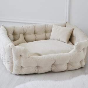May include: A cream-coloured dog bed with a tufted design and a removable pillow. The bed is made of soft, plush fabric and has a raised rim for added comfort.