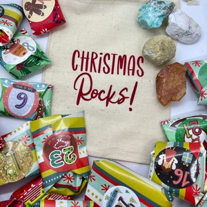 May include: A white canvas bag with the text "CHRISTMAS Rocks!" printed in red. The bag is filled with small wrapped gifts, each with a number on it, and a variety of colorful rocks.