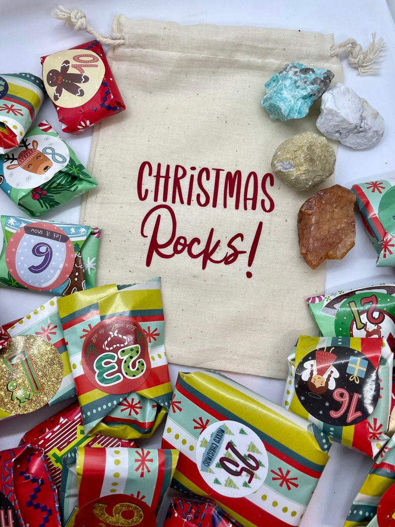 May include: A white canvas bag with the text "CHRISTMAS Rocks!" printed in red. The bag is filled with small wrapped gifts, each with a number on it, and a variety of colorful rocks.