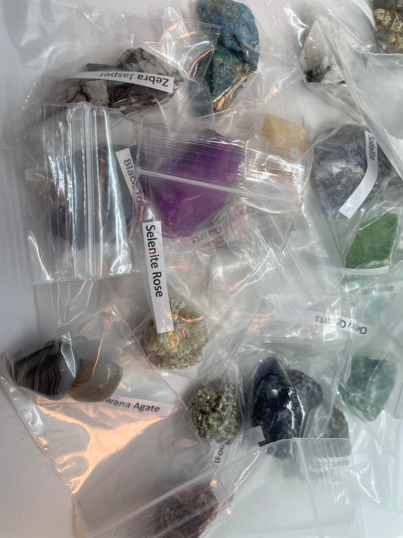 May include: A collection of various crystals and stones in individual plastic bags. The bags are labeled with the names of the stones, including Zebra Jasper, Selenite Rose, Black Tourmaline, Sodalite, Grey Quartz, and Wana Agate.