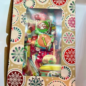 May include: A brown cardboard box with a window showing a variety of colorful Christmas advent calendar treats wrapped in festive paper. The box is decorated with a repeating pattern of snowflakes and red and green circles.