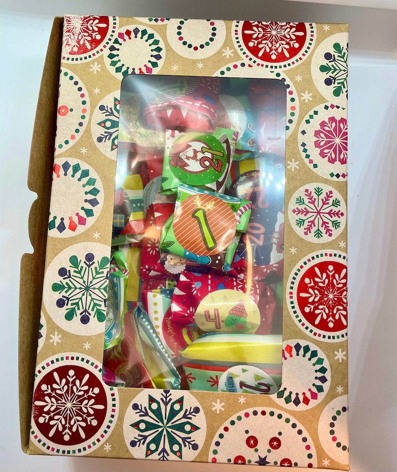May include: A brown cardboard box with a window showing a variety of colorful Christmas advent calendar treats wrapped in festive paper. The box is decorated with a repeating pattern of snowflakes and red and green circles.