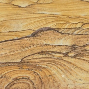 May include: A rectangular piece of polished sandstone featuring natural, flowing bands of tan, beige, and dark brown colors. The surface displays intricate, swirling, and layered patterns that resemble a desert landscape or rolling sand dunes.