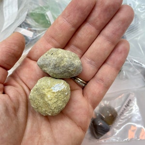 May include: Two small, round, light green and white stones with a rough texture. The stones are being held in a hand.