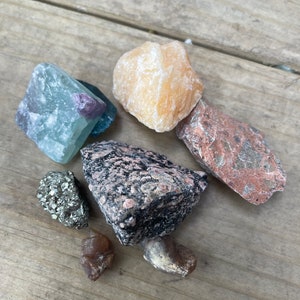 May include: A collection of seven different types of rocks and minerals, including a light blue fluorite, a yellow calcite, a dark gray granite, a red jasper, a brown agate, a gold pyrite, and a brown jasper.