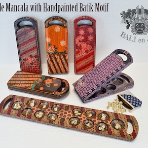 May include: A set of five wooden mancala boards with hand-painted batik designs. The boards are in various colours, including brown, orange, red, purple, and black. The boards are decorated with intricate patterns and flowers. The boards are foldable and have a handle on each side. The boards are made in Bali, Indonesia. "BALI en JAVA" is printed on the boards.