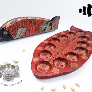 Puede incluir: Un tablero de juego de madera con forma de pez y tapa a juego, decorado con patrones pintados a mano en rojo, negro y beige. El tablero presenta catorce hendiduras circulares y se acompa&ntilde;a de peque&ntilde;as conchas blancas como fichas.