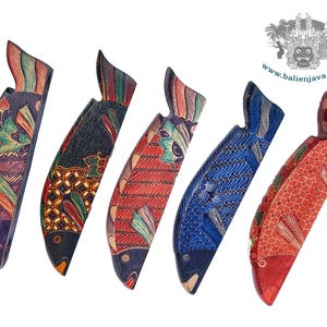 Puede incluir: Cinco figuras de peces de madera tallada dispuestas en fila, cada una con distintos patrones pintados a mano de estilo batik en morado, negro, rojo y azul. Los peces est&aacute;n orientados verticalmente con escamas y aletas detalladas. El texto dice www.balienjava.com.