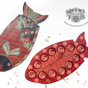 Puede incluir: Dos objetos de madera con forma de pez, uno cerrado con un patr&oacute;n batik en rojo oscuro, negro y verde azulado, y otro abierto que revela un interior rojo con hendiduras circulares. Un logotipo con el texto www.balienjava.com es visible arriba a la derecha.