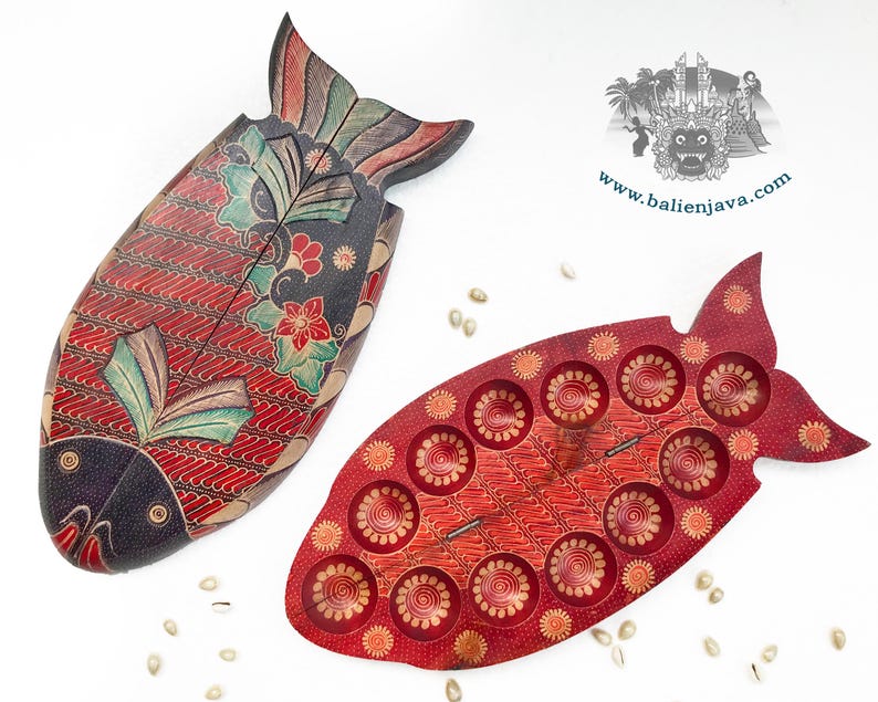Puede incluir: Dos objetos de madera con forma de pez, uno cerrado con un patr&oacute;n batik en rojo oscuro, negro y verde azulado, y otro abierto que revela un interior rojo con hendiduras circulares. Un logotipo con el texto www.balienjava.com es visible arriba a la derecha.