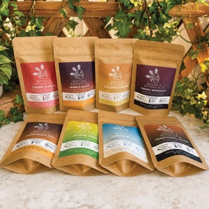 May include: An assortment of eight brown paper pouches of loose leaf tea blends. Each pouch has a colorful label with the brand name "Bathala Botanicals" and the tea type, including "Cherry Bloom" and "Mango Black".