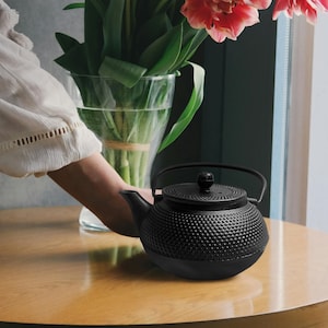 May include: A black cast iron teapot with a textured surface and a lid. The teapot has a handle and a spout. The teapot is sitting on a wooden table.