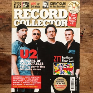 Puede incluir: Una portada de la revista "Record Collector" con U2, Johnny Cash y Elvis Presley. La portada incluye textos como "25 Years of Collectables" y "Over 200 Records Reviewed!". La revista está sobre una superficie de madera.