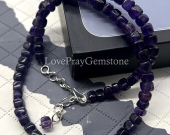 May include: A necklace made of small, square-cut amethyst beads. The beads are a deep purple color and strung together with a silver-colored chain and clasp. The text "LovePrayGemstone" is visible.