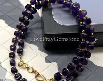 May include: A double-strand necklace featuring square, deep purple amethyst beads interspersed with small gold-colored beads. The necklace has a gold-colored clasp and the text "LovePrayGemstone" is visible.
