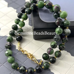 Op de afbeelding: Een dubbele ketting met ronde kralen in groen, zwart en rood. Het heeft een goudkleurige sluiting. De tekst "GemstoneBeadsBeauty" is zichtbaar. Een sieraad.