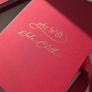 Peut inclure: Boîte rouge texturée avec couvercle assorti et ruban rouge. La boîte porte une inscription dorée sur le devant. La boîte est ouverte, révélant l'intérieur. La boîte est sur une surface blanche.