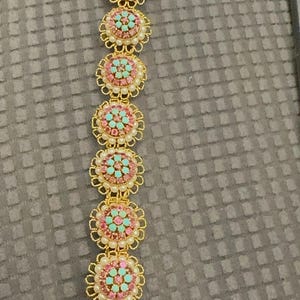Puede incluir: Un brazalete de oro con piedras rosas y turquesas dispuestas en un patrón circular. El brazalete está hecho de metal y tiene un diseño delicado.