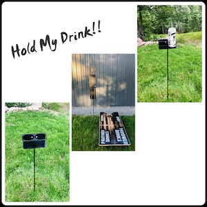 May include: Three black metal drink holders with spikes for inserting into the ground. The holders are designed to hold cans or bottles. The text "Hold My Drink!!" is written in black marker on a white background.