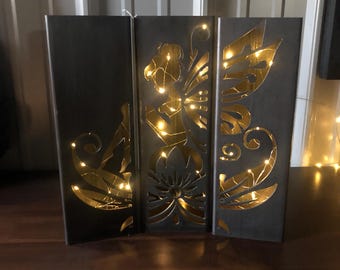 May include: A black metal fairy silhouette with cut-out details and a string of warm white lights behind it. The fairy is holding a flower and has wings. The silhouette is on a wooden surface.